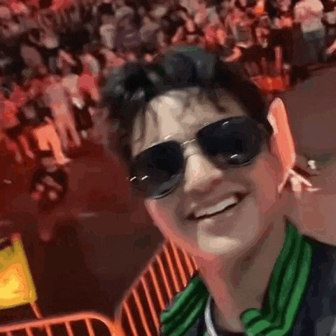 a man wearing sunglasses and a green scarf is taking a selfie in front of a crowd of people .