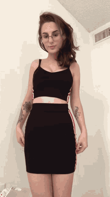 a woman wearing glasses and a black crop top is standing in front of a white wall