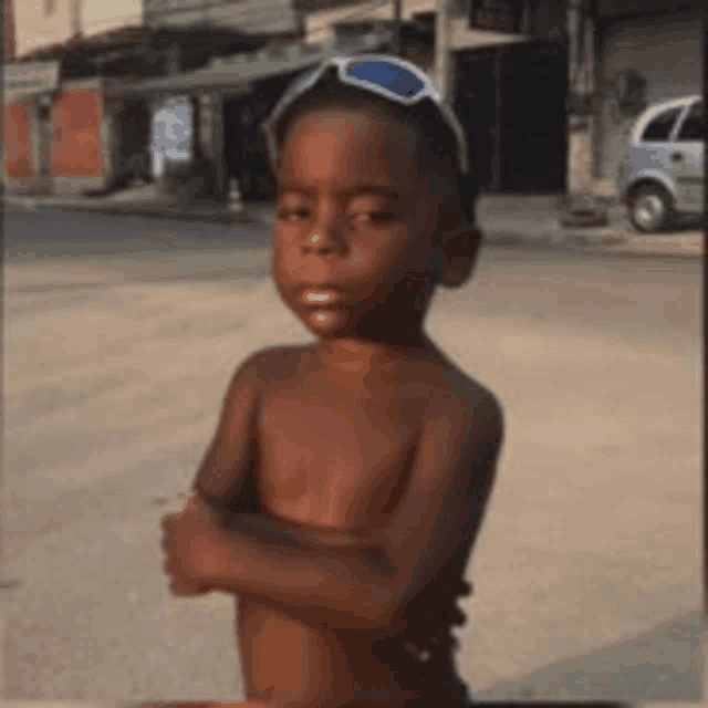 a shirtless young boy wearing sunglasses is standing with his arms crossed on a street .