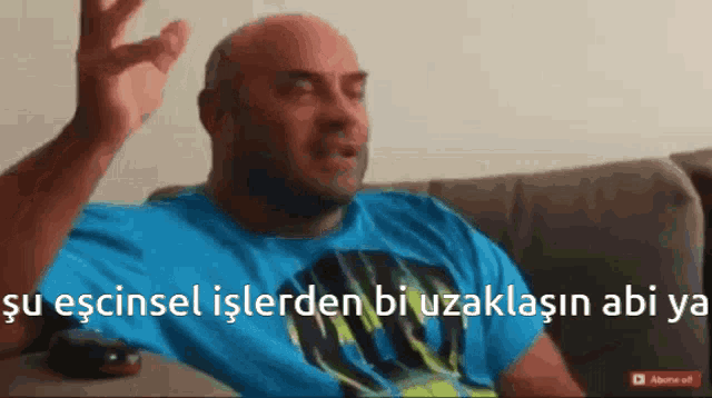 a bald man in a blue shirt is sitting on a couch with his hand up