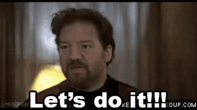 a man with a beard is standing in front of a window and says `` let 's do it ! ''