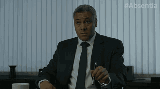 a man in a suit and tie is sitting at a desk with #absentia written on the bottom of the screen