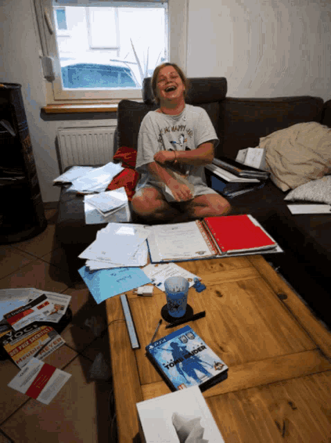 a woman is sitting on a couch laughing in front of a pile of papers and a video game called tomb raider