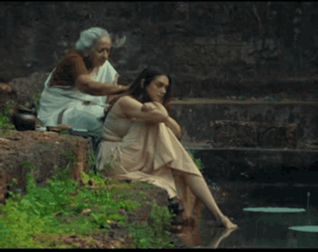a woman is sitting next to another woman who is sitting on a rock near a body of water