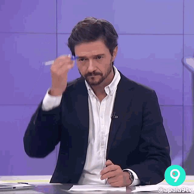 a man in a suit is sitting at a desk with a pen in his hand in front of a sign that says 9