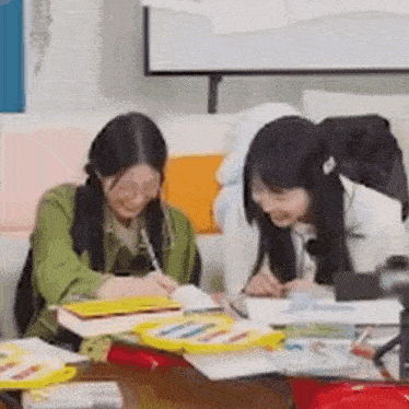 two girls are sitting at a table writing in notebooks and smiling at each other .