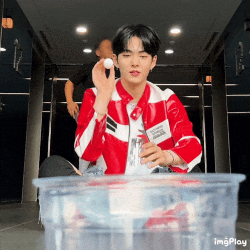 a man in a red jacket is holding a white ball in his hand while sitting next to a plastic container of water
