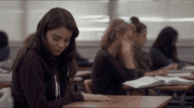 a girl is sitting at a desk in a classroom with other students while reading a book .