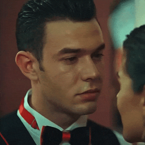 a man in a tuxedo and bow tie looks at a woman in a white shirt