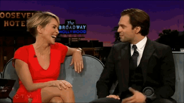 a woman in a red dress sits next to a man in a suit in front of a sign for the broadway hollywood