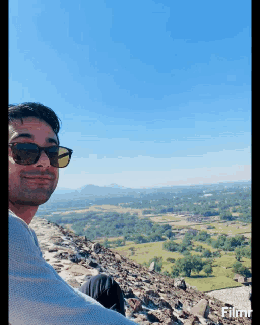 a man wearing sunglasses sits on top of a rocky hill overlooking a landscape