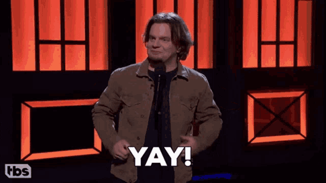 a man in a brown jacket stands in front of a microphone saying yay
