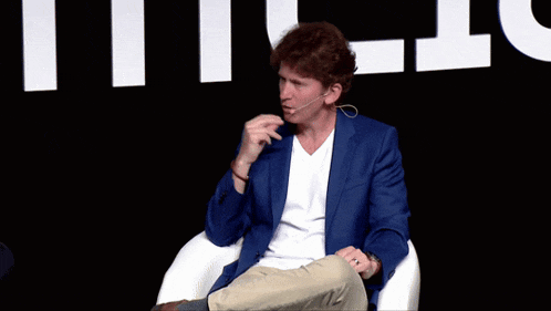 a man wearing a blue jacket and a white shirt is sitting in front of a sign that says ' t '