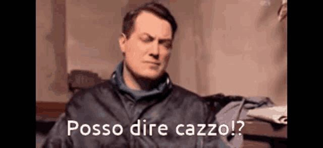 a man in a black jacket is sitting on a bed with the words posso dire cazzo written on the screen behind him .