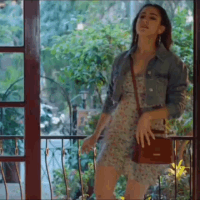 a woman in a floral dress and denim jacket is standing in front of a window .