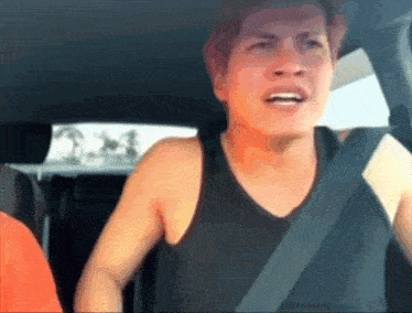 a man wearing a black tank top is sitting in a car with a seat belt around his neck