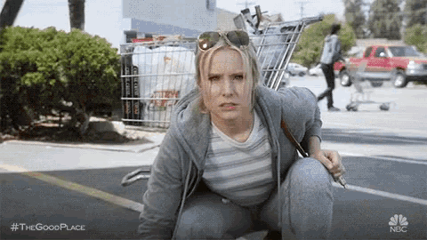 a woman is squatting down in a parking lot next to a shopping cart filled with groceries .