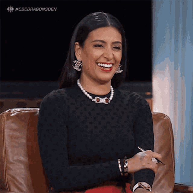 a woman wearing a pearl necklace and chanel earrings laughs while sitting in a chair