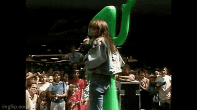 a woman is singing into a microphone while standing next to a green inflatable gummy man .