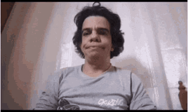 a man wearing a gray shirt is making a funny face while sitting on a bed .