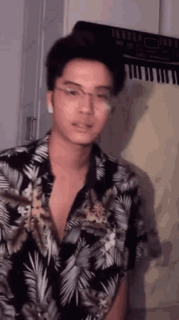 a young man wearing glasses and a hawaiian shirt is standing in front of a piano .