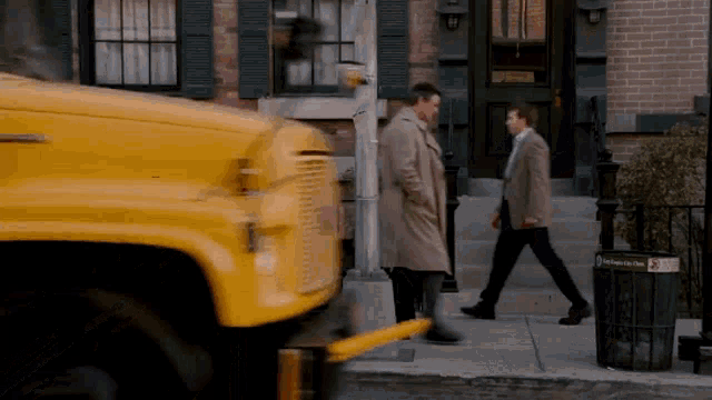 a man in a trench coat is walking down a sidewalk next to a yellow bus