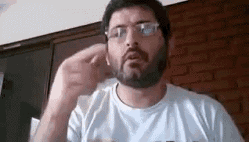 a man with a beard and glasses is making a funny face while sitting in front of a brick wall .