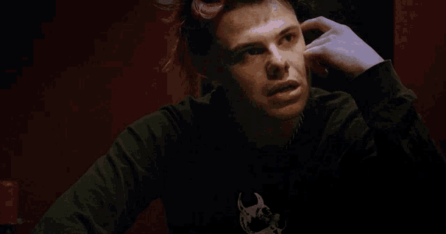a young man with pink hair is sitting in a dark room with his hand on his head .