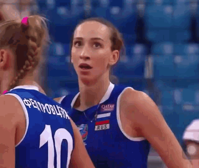 two female volleyball players are talking to each other in a stadium . one of the players has the number 10 on her jersey