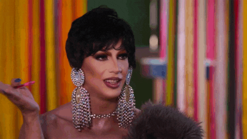 a drag queen wearing earrings and a necklace is standing in front of a colorful background .