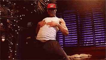 a man wearing a red hat and a white tank top is dancing in front of a christmas tree .