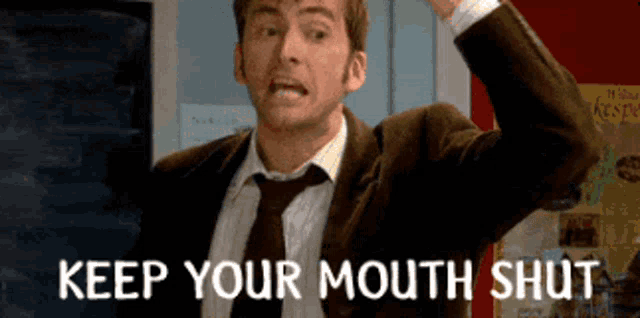 a man in a suit and tie says " keep your mouth shut " in front of a chalkboard