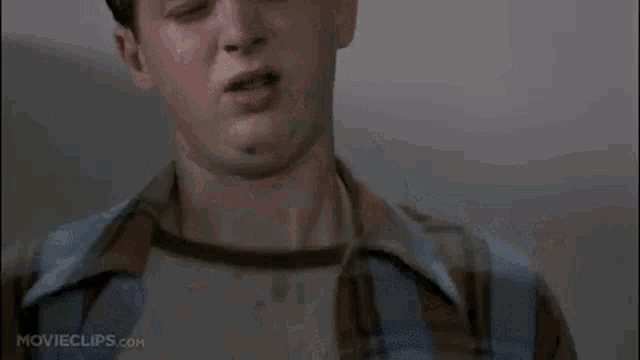 a man in a plaid shirt is sitting in a public restroom with a roll of toilet paper .