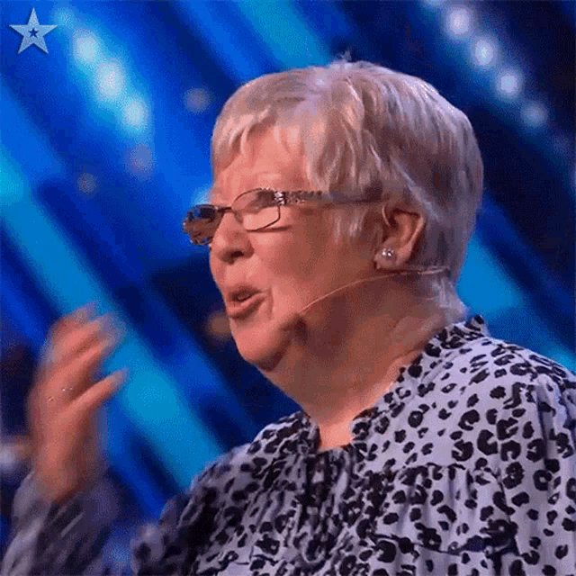 an elderly woman wearing glasses and a leopard print shirt is speaking into a microphone on a stage .