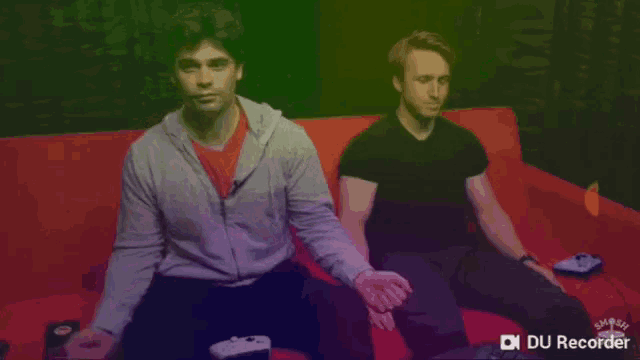 two men sitting on a red couch with a du recorder on the bottom left