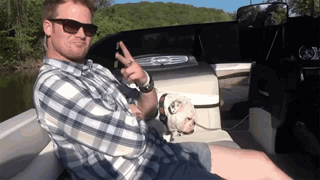 a man in a plaid shirt is sitting on a boat with a dog and giving a peace sign