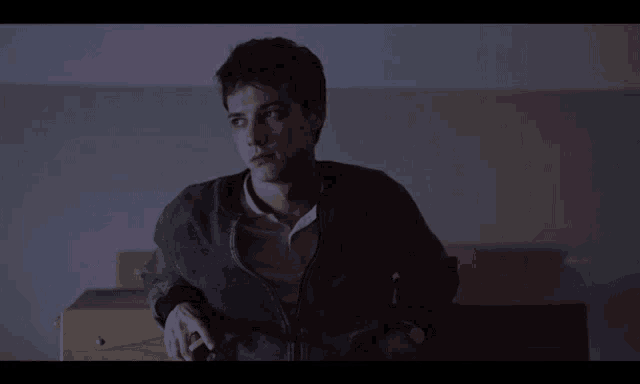 a man in a gray jacket sits in a dark room looking at something