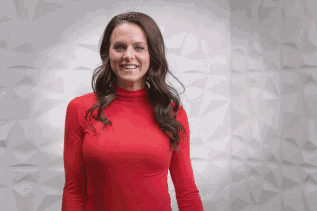 a woman in a red turtleneck sweater smiles in front of a white wall