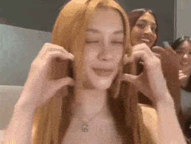 a woman with long red hair and a necklace is making a heart shape with her hands .