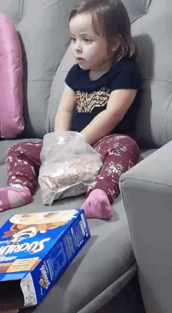 a little girl sits on a couch with a box of cheerios in front of her