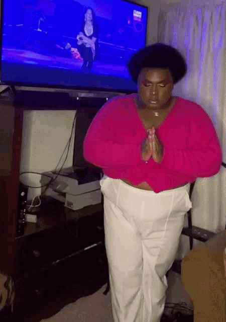 a woman in a pink sweater and white pants is praying in front of a tv
