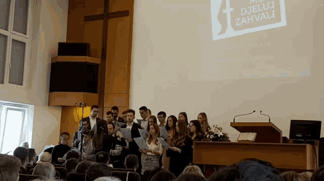 a group of people are singing in front of a large screen that says djeluj zahvali