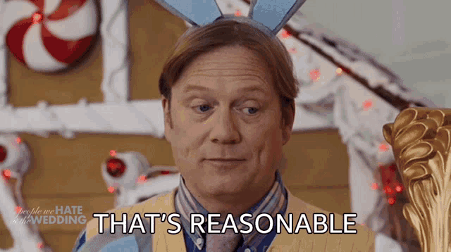 a man wearing bunny ears says that 's reasonable in front of a gingerbread house