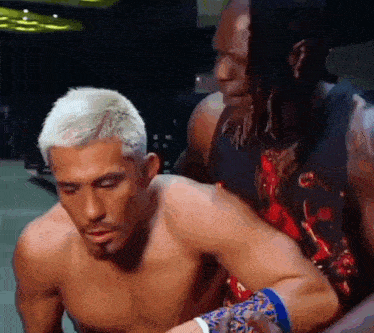 a shirtless wrestler is being helped by another wrestler in a wrestling ring .