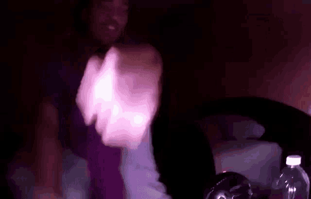 a man in a purple shirt is dancing in a dark room with a bottle of water in the background .