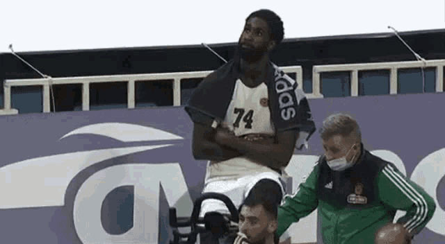 a basketball player is sitting on a wheelchair with his arms crossed while a man in a mask looks on .