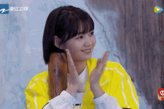 a woman in a yellow jacket is clapping her hands in front of a tv screen that says ' a ' on it