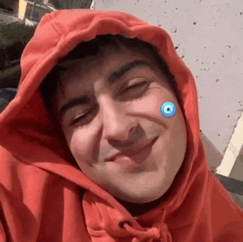 a man wearing a red hoodie with a blue evil eye on his face is smiling .