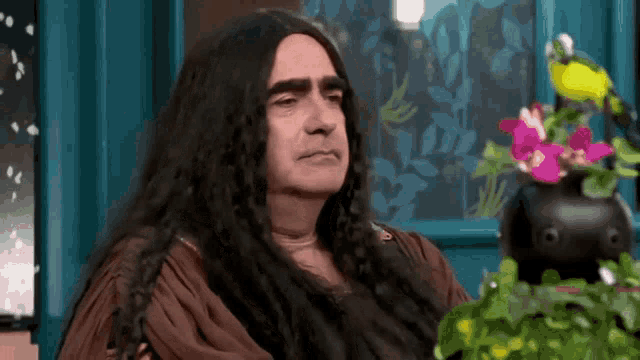 a man with long hair is sitting in front of a window with flowers in it .