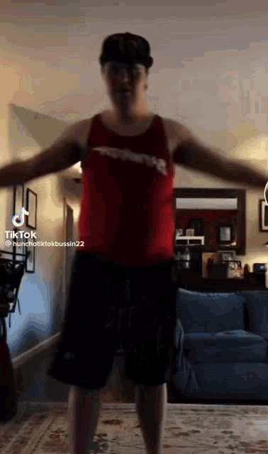 a man in a red tank top and black shorts is jumping in a living room with his arms outstretched .